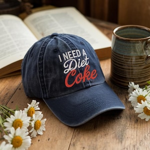 May include: A navy blue baseball cap with the text "I NEED A Diet Coke" in white and red lettering. The cap is sitting on a wooden surface with a book, a mug, and daisies in the background.