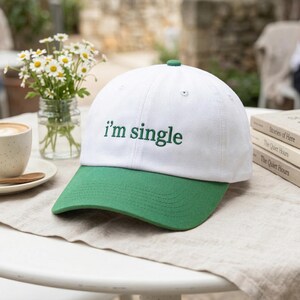 May include: White and green baseball cap with the text "i'm single" embroidered in green. The cap has a green brim and a green button on top. The hat is sitting on a white table.