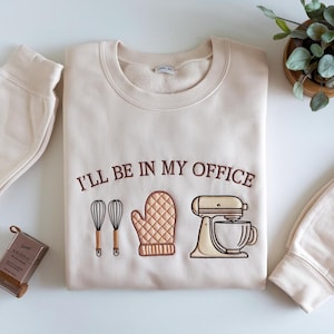 May include: Cream-colored sweatshirt with the embroidered phrase "I'LL BE IN MY OFFICE" above illustrations of a whisk, oven mitt, and stand mixer. The sweatshirt is folded, with a matching beige sweatpant cuff visible.
