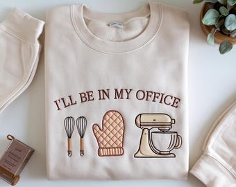 May include: Cream-colored sweatshirt with the embroidered phrase "I'LL BE IN MY OFFICE" above illustrations of a whisk, oven mitt, and stand mixer. The sweatshirt is folded, with a matching beige sweatpant cuff visible.