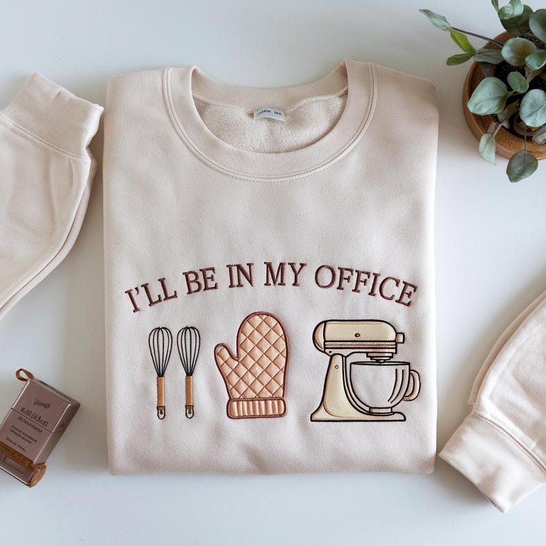 May include: Cream-colored sweatshirt with the embroidered phrase "I'LL BE IN MY OFFICE" above illustrations of a whisk, oven mitt, and stand mixer. The sweatshirt is folded, with a matching beige sweatpant cuff visible.