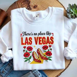 May include: White sweatshirt with the text "There's no place like LAS VEGAS" in red and yellow. The design includes red flowers and a pair of red high heels. The sweatshirt is displayed on a wooden surface.