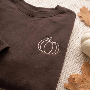 May include: A dark brown crewneck sweatshirt featuring a simple, light-colored embroidered outline of a pumpkin on the left chest area. The garment has a ribbed collar and long sleeves, showcasing a clean, minimalist design with a single line art motif.
