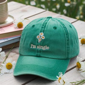 May include: A green baseball cap with the embroidered text "I'm single" and a small floral design. The cap is made of a soft material and has a curved brim. The hat is a casual accessory.