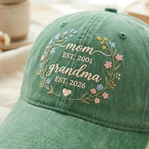 May include: Green baseball cap with embroidered floral wreath design. The text reads "mom EST. 2001" and "grandma EST. 2026" with a small heart. A stylish accessory for mothers and grandmothers.