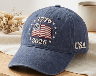 May include: A blue baseball cap with the text "1776" and "2026" embroidered above and below an American flag design. The word "USA" is embroidered on the side. The cap is sitting on a wooden surface.