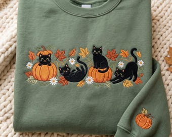 Op de afbeelding: Een olijfgroene sweater met ronde hals en een geborduurd ontwerp van drie zwarte katten, oranje pompoenen, herfstbladeren en witte madeliefjes. Een kleine bijpassende pompoen is geborduurd op de mouwboord van het kledingstuk.