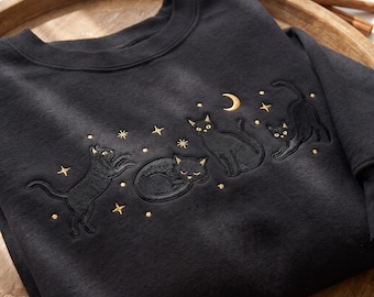 Könnte beinhalten: Ein schwarzes Sweatshirt mit Rundhalsausschnitt und goldener Stickerei, die drei Katzen in verschiedenen Posen zeigt, darunter eine spielende, eine schlafende und eine sich streckende Katze, umgeben von kleinen goldenen Sternen und einem Halbmond.