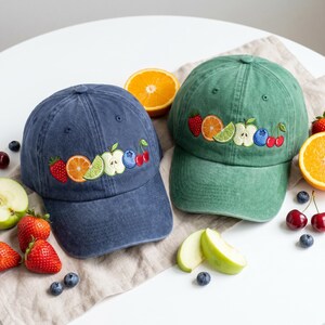 May include: Two baseball caps, one navy blue and one sage green, each featuring an embroidered row of colorful fruit including a strawberry, orange slice, lime wedge, apple slice, blueberry, and cherries on the front panel of the crown.