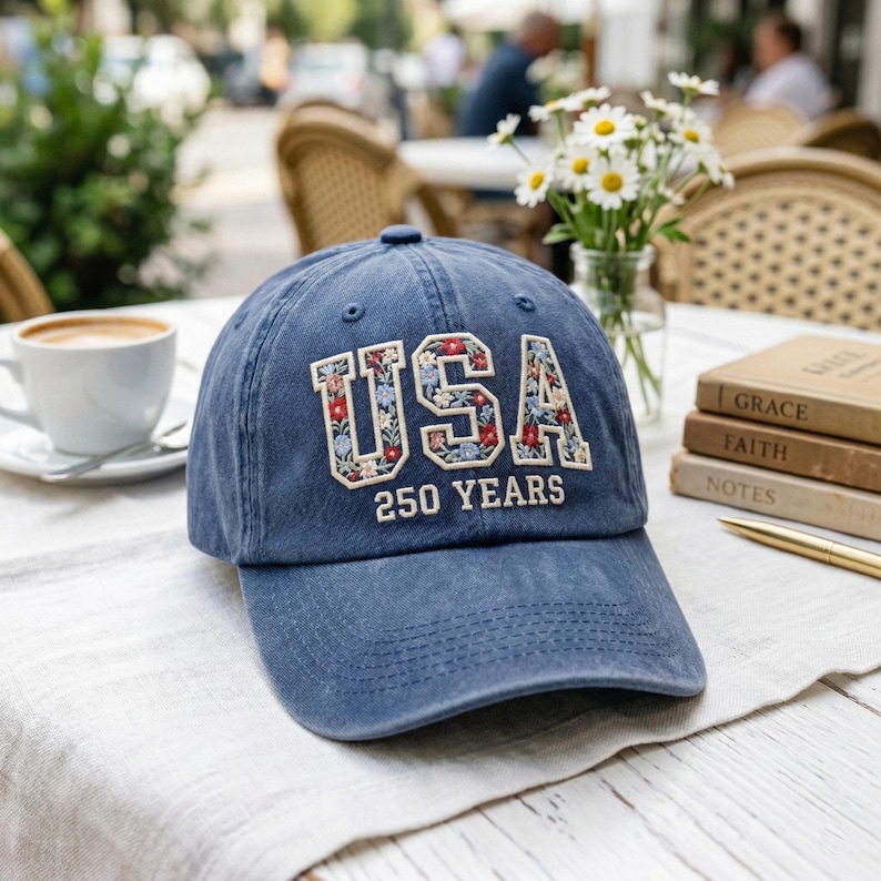 Puede incluir: Gorra de b&eacute;isbol azul marino con la palabra "USA" bordada con letras florales. El texto "250 YEARS" est&aacute; debajo. La gorra est&aacute; sobre una superficie blanca, con una taza de caf&eacute; y un jarr&oacute;n de flores en el fondo.