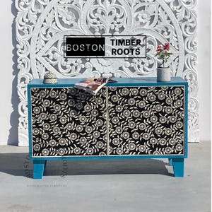 May include: A blue cabinet with two doors, each featuring a black and white floral design. The cabinet is made of wood and has a modern style. The cabinet is sitting on a white tiled floor. The text "BOSTON TIMBER ROOTS" is visible on the cabinet.