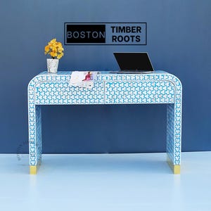 Puede incluir: Una consola de color blanco y azul con dos cajones y detalles dorados. La mesa está hecha de incrustaciones de hueso y tiene un diseño moderno. La mesa mide 122 cm de largo y 46 cm de profundidad. La mesa está sobre un suelo de color azul claro frente a una pared azul. El texto "BOSTON TIMBER ROOTS" es visible en la pared.