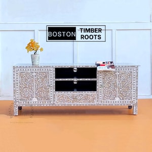 May include: A gray and white inlaid wood TV stand with two doors, two shelves, and a drawer. The stand has a floral design and the words "BOSTON TIMBER ROOTS" are printed on the top.