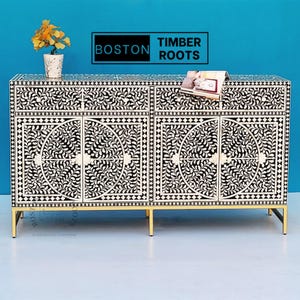 Puede incluir: Un aparador de incrustaciones de hueso en blanco y negro con patas de metal dorado. El aparador tiene cuatro puertas y dos cajones. Las puertas y los cajones presentan un diseño floral. El aparador está etiquetado como "BOSTON TIMBER ROOTS".
