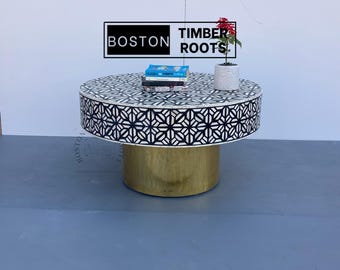 May include: A round, black and white bone inlay coffee table with a gold metal base. The table top features a geometric pattern. The table is sitting on a gray surface. BOSTON TIMBER ROOTS is printed on a white rectangle behind the table.