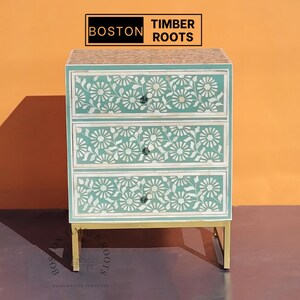 May include: A three-drawer chest with a floral design. The chest is teal with white floral inlays and gold-colored legs. The top of the chest has a floral pattern. The brand name "BOSTON TIMBER ROOTS" is at the top.