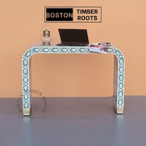 May include: A white console table with a blue and white geometric pattern inlay. The table has a curved top and two legs. A laptop, a magazine, and a lantern are on the table.