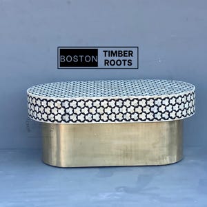 Op de afbeelding: Ovale salontafel met een geometrisch patroon in zwart en wit op de bovenkant en een gouden metalen onderstel. De tafel is gelabeld met "Boston Timber Roots".