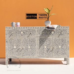 Puede incluir: Un aparador gris con diseño floral y detalles de estrella. El mueble tiene cuatro puertas y dos cajones. El texto "BOSTON TIMBER ROOTS" se muestra en la parte superior. Una pequeña planta en maceta blanca encima.