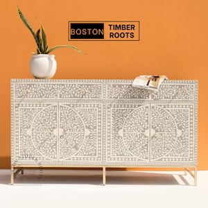 May include: A gray and white cabinet with a floral design, featuring four doors and two drawers. The cabinet has gold-colored legs and handles. A white pot with a green plant sits on top, along with a magazine. The text "BOSTON TIMBER ROOTS" is displayed.