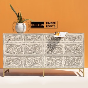 May include: A gray and white dresser with six drawers, adorned with a leaf pattern. The dresser has gold-colored legs and drawer pulls. A potted plant and a book rest on top. The brand name "BOSTON TIMBER ROOTS" is visible.