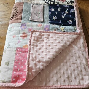 May include: A pink and white patchwork baby quilt with a soft, pink, textured backing. The quilt is made from a variety of fabrics, including floral prints, polka dots, and solid colors. The quilt is folded in half and has a pink binding around the edges.