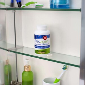 May include: A white bottle of Prevagen dietary supplement with blue and green labels, displayed on a glass shelf inside a medicine cabinet. The bottle's label reads "Prevagen Improves Memory Regular Strength". A single capsule is next to the bottle.