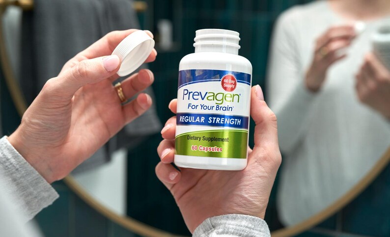 May include: A white bottle of "Prevagen For Your Brain" dietary supplement, held in hands. The bottle has a blue and white label with the words "Regular Strength" and "60 Capsules". The lid is being removed.