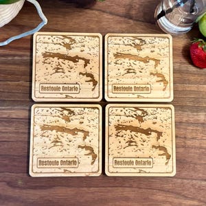 Peut inclure: Quatre dessous de verre carrés en bois avec un motif de carte et le texte "Restoule Ontario" gravé sur chacun. Les dessous de verre sont brun clair et reposent sur une surface en bois brun foncé. Une fraise, une poire et une bouteille en verre sont également visibles.