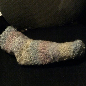 May include: A handmade sock in shades of blue, pink, and yellow. The sock is made of a textured, fuzzy yarn. The sock is lying on a black surface. The sock appears to be hand-knitted.