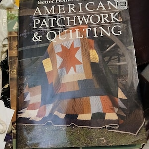 Pode incluir: Um livro intitulado "American Patchwork & Quilting" da Better Homes and Gardens. A capa apresenta um edredom de patchwork colorido com um desenho de estrela. O livro está rodeado por outros livros e uma superfície castanha escura.