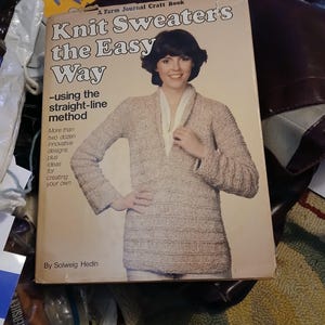 May include: A vintage book cover titled "Knit Sweaters the Easy Way" by Solweig Hedin. The cover features a woman wearing a beige knit sweater. The book's title and author are prominently displayed.