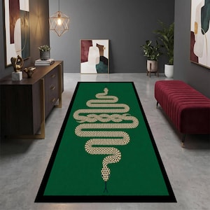 Puede incluir: Una alfombra rectangular con fondo verde intenso y borde negro. Centrada en la alfombra hay una gran ilustración de serpiente estilizada en color beige claro con un patrón de escamas geométricas, representada en una posición horizontal sinuosa.