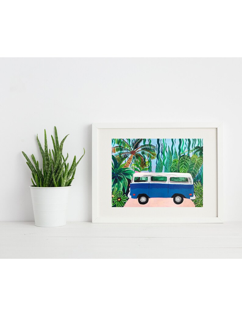 May include: A framed art print featuring a blue and white vintage van parked on a pink road, set against a backdrop of lush green tropical foliage. The artwork is in a white frame, evoking a sense of travel and adventure.