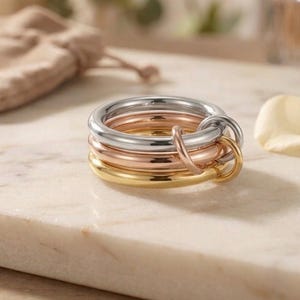 Peut inclure: Un ensemble de trois anneaux entrelacés aux tons argent, or rose et or jaune. Les anneaux présentent un design lisse, poli et arrondi, et sont reliés entre eux par un petit anneau métallique assorti qui unit les trois bandes distinctes.