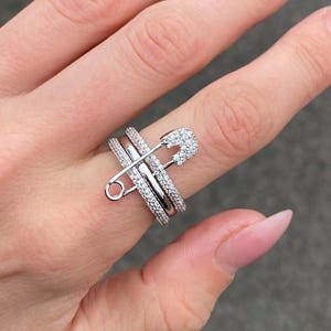 Peut inclure: Une bague argentée arborant un motif d'épingle à nourrice ornée de petites pierres claires et scintillantes. L'anneau se compose de plusieurs rangées de pierres serties pavé qui entourent le doigt, créant un aspect superposé.