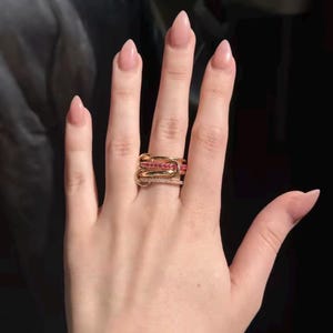 Peut inclure: Une pile de quatre bagues en or portées sur un doigt. L'ensemble comprend deux anneaux en or poli, un anneau orné d'une rangée de petites pierres roses et un anneau incrusté d'une ligne de minuscules pierres blanches, disposés en couches.