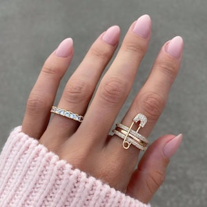 Peut inclure: Une main portant trois bagues en or. L'une est une alliance sertie de petites pierres transparentes. Une autre est un design superposé avec une forme d'épingle à nourrice incrustée de pierres transparentes. La troisième est une alliance en or avec des pierres transparentes.