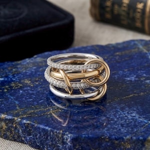 Peut inclure: Une bague multi-anneaux composée de cercles entrelacés aux tons argentés et dorés. Deux des anneaux sont incrustés de petites pierres claires serties pavé, tandis que les autres sont en métal lisse et plein, créant un aspect tissé superposé.