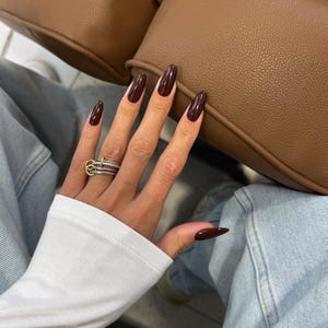 Peut inclure: Une bague multi-anneaux composée de métal argenté et d'un petit accent circulaire doré. La bague est portée sur un doigt aux ongles longs en forme d'amande, vernis d'une couleur brun chocolat foncé brillant.