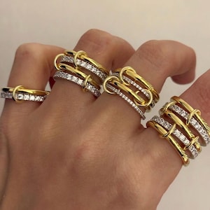 Peut inclure: Une collection de bagues dorées composées de plusieurs anneaux, dont certains sont incrustés de petites pierres claires et scintillantes. Les bagues présentent des boucles entrelacées et des structures superposées sur les doigts.