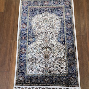 Könnte beinhalten: Ein rechteckiger Gebetsteppich mit einem zentralen bogenförmigen Design. Der Teppich hat ein weißes Feld mit komplizierten floralen Mustern in Blau-, Rot- und Goldtönen. Ein dunkelblauer Rand mit floralen Motiven umgibt das zentrale Design. Der Teppich hat gefranste Kanten.