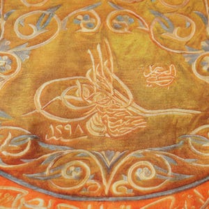 Pu&ograve; includere: Primo piano di un tessuto circolare di colore arancione e oro con intricati disegni floreali bianchi e calligrafia araba. Il tessuto ha un bordo blu e la data 1291 &egrave; visibile al centro.