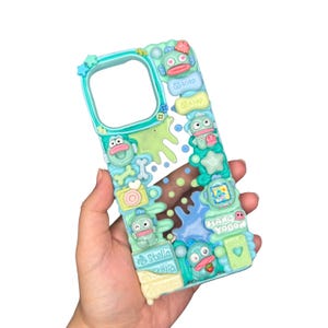 May include: A teal phone case with a whimsical design featuring cartoon characters, stars, and the word "soap". The case has a textured, 3D appearance with various shapes and colors, including green, blue, and yellow accents.