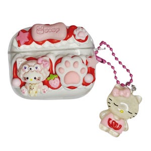 Può includere: Custodia trasparente per auricolari con design rosa e bianco. La custodia presenta un gatto dei cartoni animati, impronte di zampe e una fragola. Un ciondolo a forma di gatto abbinato è appeso a una catena rosa. La parola "soap" è in alto.