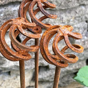 May include: Three metal garden stakes featuring spherical tops constructed from spiraling, rusted iron bands. Each sphere is attached to a straight, thin metal rod, displaying a weathered, oxidised orange-brown texture throughout the entire structure.