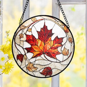 May include: A round stained glass window hanging with a black metal frame and chain. The design features a large red, orange, and yellow maple leaf in the center, surrounded by smaller brown leaves and white background.
