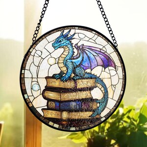 May include: Round stained glass window art featuring a blue dragon perched atop a stack of antique books. The dragon has purple wings and is set against a white and beige background. A black chain is attached for hanging.