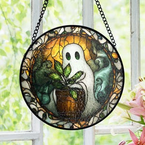 May include: Round stained glass window art featuring a white ghost holding a potted plant. The artwork has a black metal frame and chain for hanging. The ghost has two green eyes and is surrounded by a colorful background of yellow, green, and blue.
