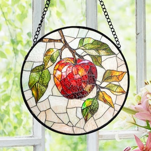 May include: Round stained glass window art featuring a red apple on a branch with green and yellow leaves. The artwork is framed in black and hangs from a black chain. The background is a mosaic of off-white glass.